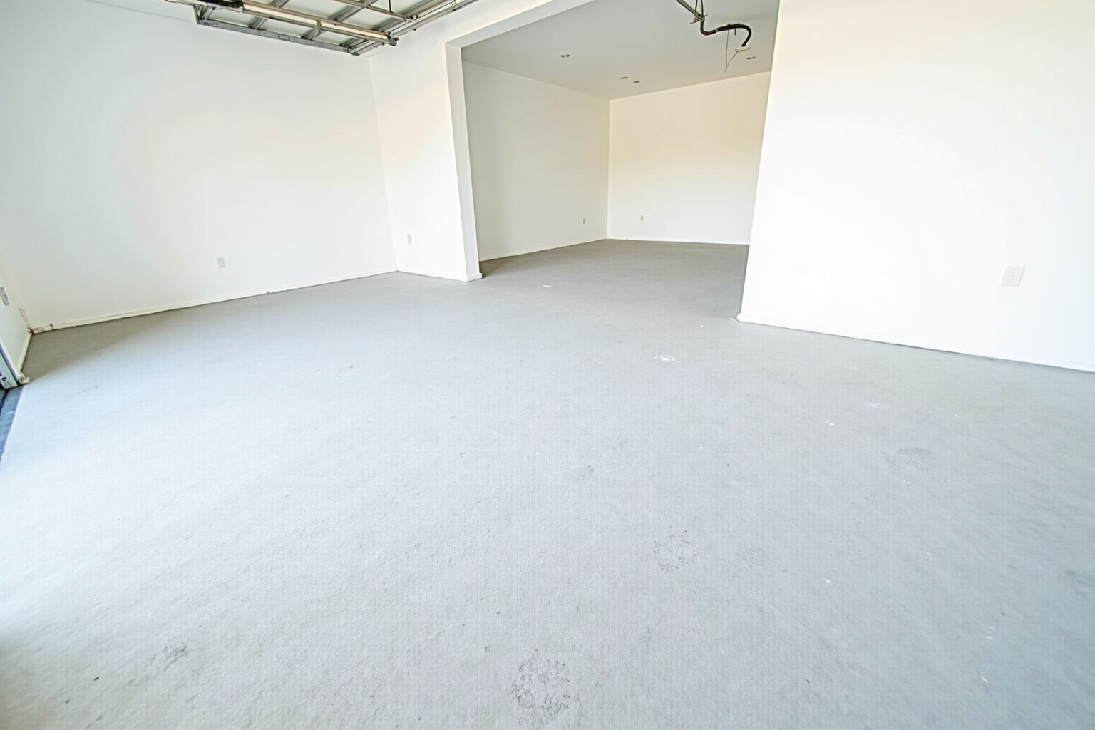 A heavy-duty industrial grinder smooths a dusty gray floor, creating a cloud of fine concrete particles. Strong overhead lighting casts sharp shadows, highlighting the abrasive texture of the unfinished floor surface.
